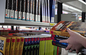 Das Bild zeigt ein Regal in der Uni-Bibliothek. Darauf stehen Bücher. Im Vordergrund hält eine Hand einen Stapel Bücher.