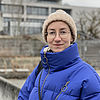 Eine junge Frau mit Brille, Wollmütze und blauer Winterjacke steht vor einem Hochschulgebäude