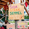 Ein Plakat, bestehend aus einem braunen Pappkarton, mit der Aufschrift:  SOJA STEAK SEMMEL, Spendenempfehlung: 3 - 5 €; darunter ein kleineres Palakt mit der Aufschrift: alles vegan