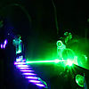 In einer dunklen Umgebung trifft geht ein grüner Lichtstrahl von rechts nach links auf eine Problenlösung in einer Phiole, die einem unterbrochenen violetten Laserstrahl ausgesetzt ist.