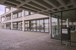 Eingangsbereich eines Gebäudekomplexes in Sichtbetonweise; vor der Glasfront ist ein großformatiger Briefkasten zu sehen.