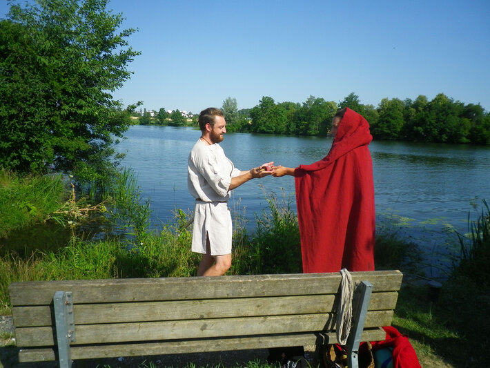Eine mit einem roten Kapuzenumhang verhüllte Person übergibt einem mit einer kurzen Tunika bekleideten Mann ein Feldzeichen. (öffnet Vergrößerung des Bildes)