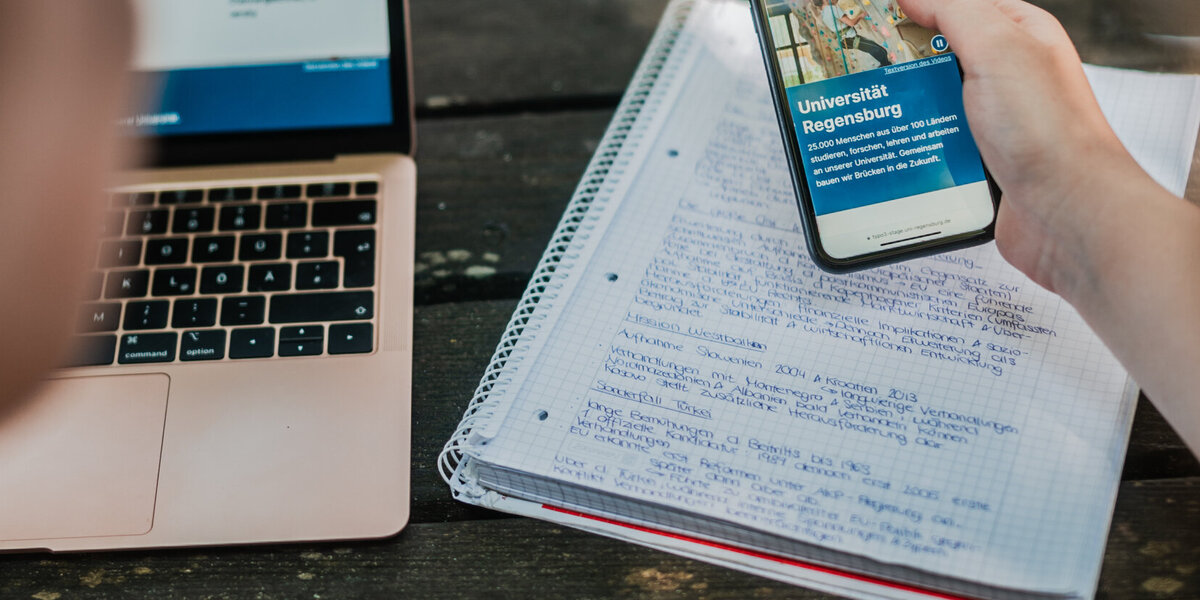 Zwei Studierende blicken gemeinsam auf das Display eines Smartphones. Im Hintergrund sind ein Laptop und ein beschriebener Notizblock zu sehen.