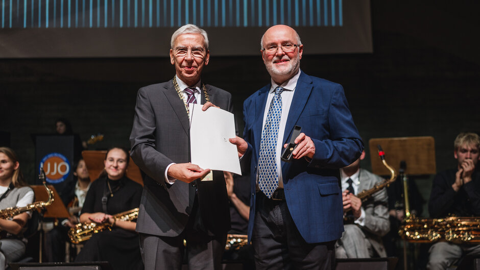 Zwei Herren in dunklen Anzügen stehen auf der Bühne, hinter sich Musikerinnen und Musiker des Uni-Jazzorchesters mit Blasinstrumenten. Sie halten gemeinsam eine Urkundenmappe hoch, der rechte Herr hat ein Futteral mit einer Anstecknadel in der Hand.