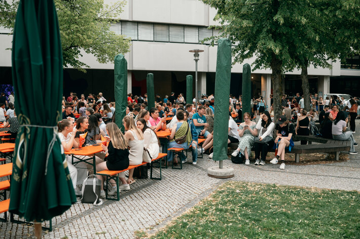 Besucherinnen und Besucher des Uni-Sommerfests sitzen unter Bäumen auf Biertischgarnituren vor dem PT-Gebäude. (öffnet Vergrößerung des Bildes)