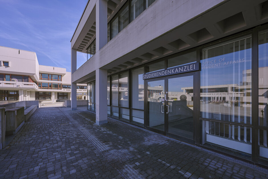Der Eingang in die Studierendenkanzlei der Universität Regensburg; in den Glasscheiben der Fenster und der Eingangstür spiegelt sich das Forum der Universität.