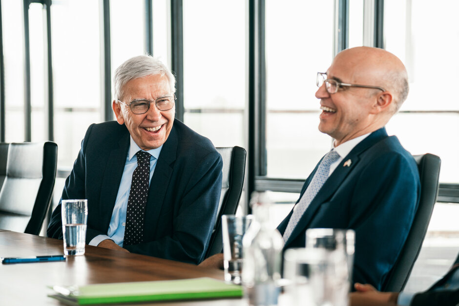 Zwei Männer im Anzug sitzen an einem Konferenztisch und lachen herzlich.