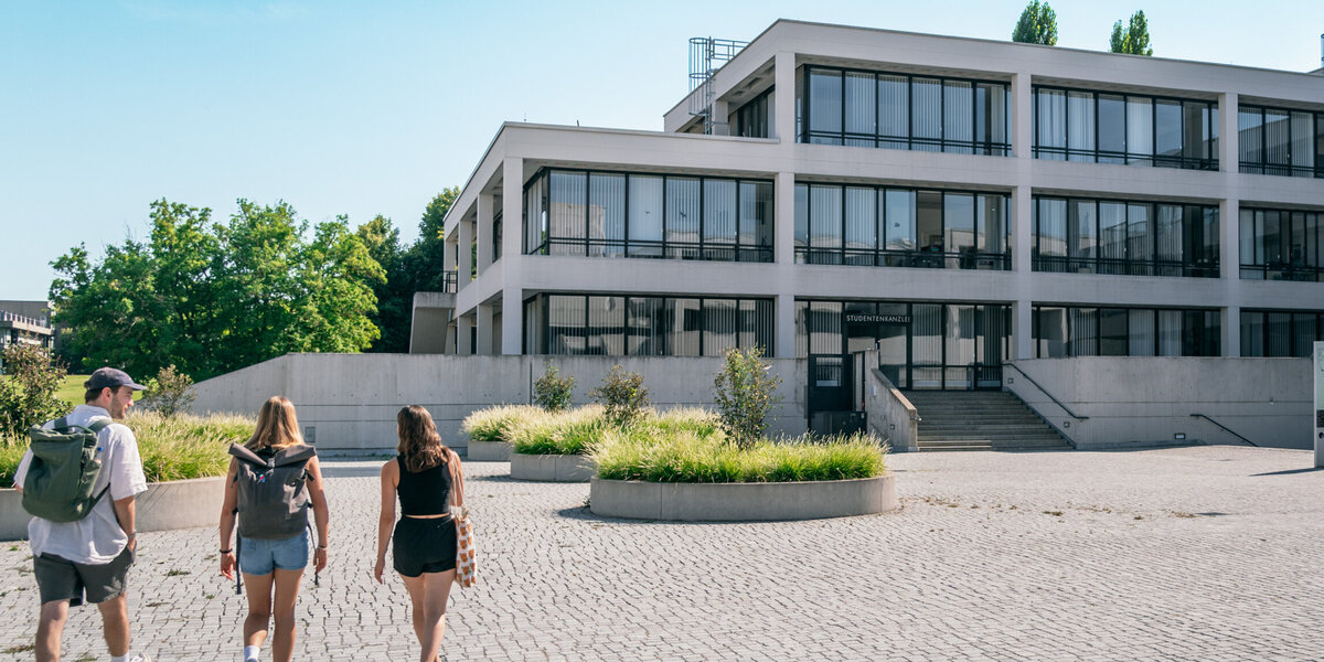 Drei Studierende gehen über das Forum der Universität Regensburg Richtung Studierendenkanzlei.