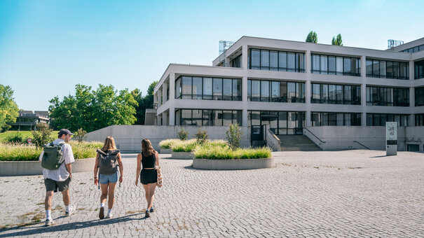 Drei Studierende gehen über das Forum der Universität Regensburg Richtung Studierendenkanzlei.