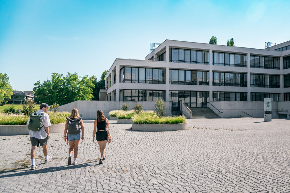 Drei Studierende gehen über das Forum der Universität Regensburg Richtung Studierendenkanzlei.