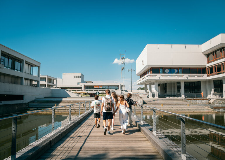 Studierende gehen auf der Brücke über den Uni-See in Richtung Zentrales Hörsaalgebäude der Universität Regensburg.