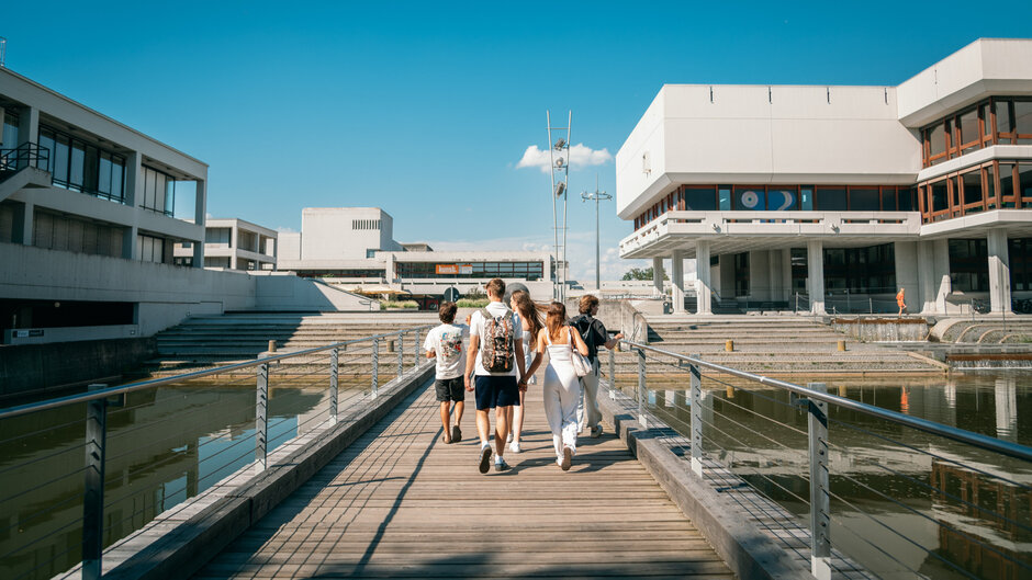 Studierende gehen auf der Brücke über den Uni-See in Richtung Zentrales Hörsaalgebäude der Universität Regensburg.