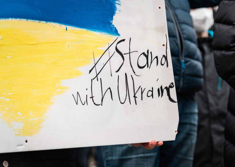 Bei einer Demonstration wird ein Plakat mit gelber und blauer Farbe sowie dem Schriftzug "Stand with Ukraine" gezeigt.