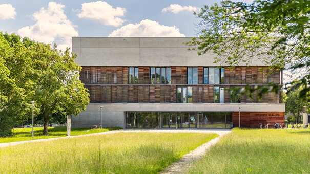 Ein Hochschulgebäude mit Beton- und Holzfassade steht in einer parkähnlichen Umgebung umgeben von grüner Wiese, grünen Bäumen. Die Sonne scheint, der Himmel ist blau mit vereinzelten weißen Wolken.