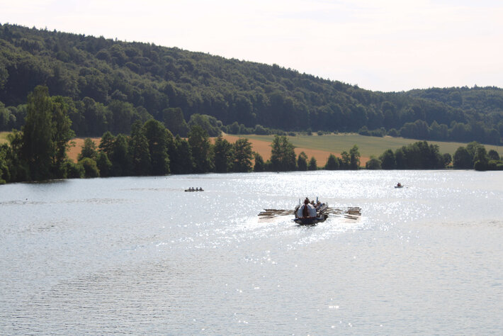 In einiger Entfernen fahren ein kleines Ruderboot und ein großen Ruderboot auf einem breiten Fluss. Im Hintergrund sind Felder und bewaldete Hügel zu sehen. (öffnet Vergrößerung des Bildes)