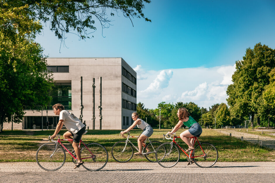 Drei Studierende fahren mit ihren Fahrrädern über den sommerlichen Campus der Universität Regensburg.