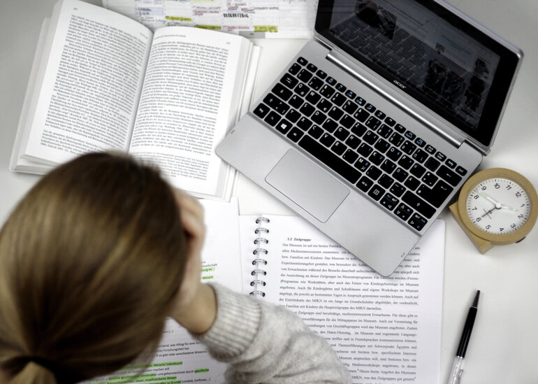 Eine Studentin sitzt an einem Schreibtisch, auf dem ein Laptop, ein Wecker, auf aufgeschlagenes Buch und ein Vorlesungsskript liegen.