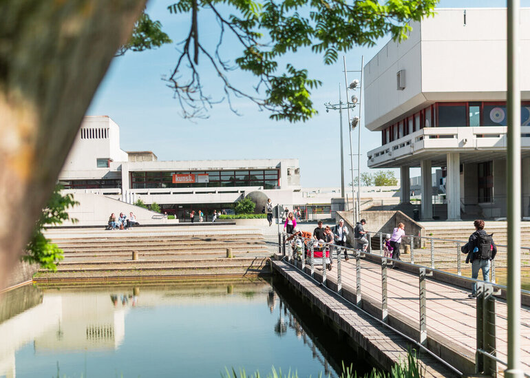 Der See der Uni Regensburg mit einer Brücke, auf der einige Menschen zu sehen sind. Im Hintergrund sieht man das Audimax.