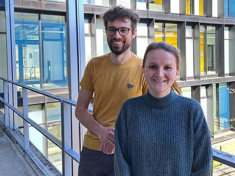 Ein Mann mit Brille und Bart und eine Frau mit zusammengebundenen Haaren auf einem Balkon vor einem Büro- oder Seminargebäude mit vielen Fenstern.