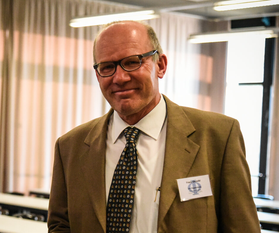 A 60-year-old man in a brown suit smiles directly into the camera. (opens enlarged image)