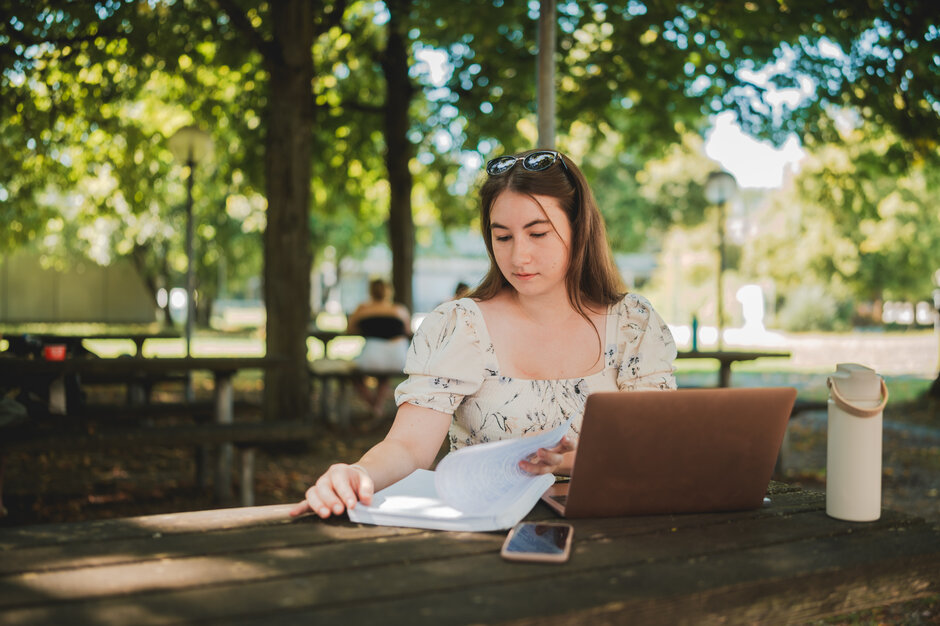 Eine Studentin sitzt mit Laptop und Studienunterlagen im Freisitz der Chemie-Cafete an der Universität Regensburg
