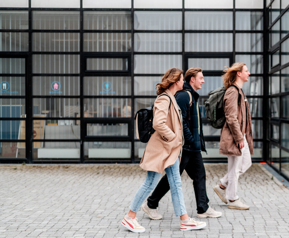 Drei Studierende gehen an der Fensterfront des Studierendenhauses an der der Universität Regensburb vorbei.