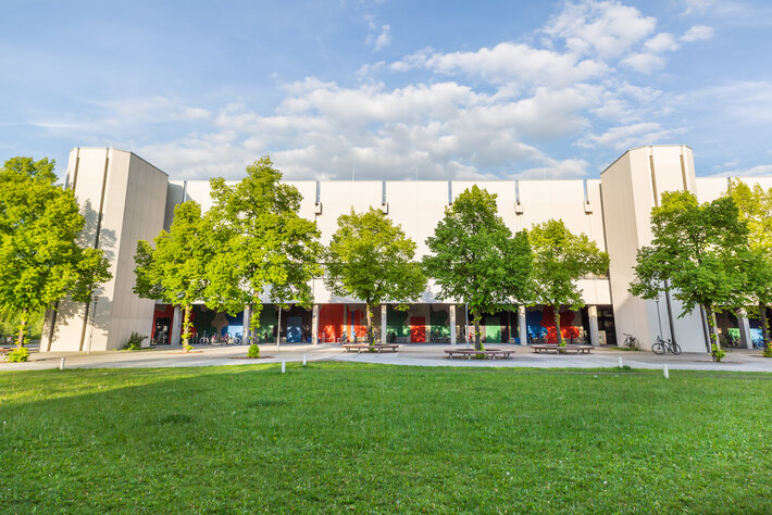 Blick über eine Wiese und Bäume auf den Gebäudeteil Philosophie/Theologie am Campus der Universität Regensburg (öffnet Vergrößerung des Bildes)
