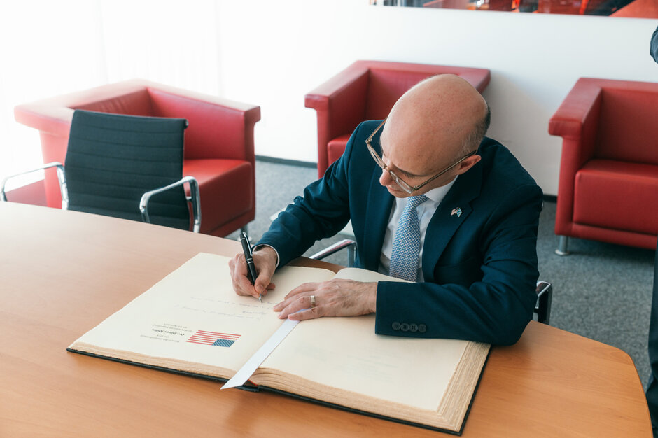 Ein Mann im Anzug schreibt mit Füller in ein großformatiges Buch. Über seinem Text wurde ein Stück Papier mit der aufgedruckten US-amerikanische Flagge eingeklebt.