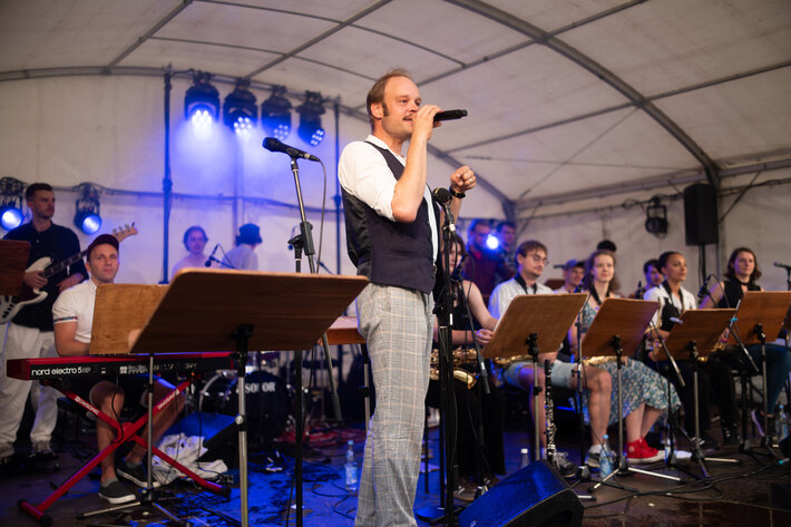 Ein Mann mit heller Hose, weißem Hemd und schwarzer Weste steht auf der Bühne und spricht in ein Mikrofon. Im Hintergrund sind die Musikerinnen und Musiker des Uni-Jazzorchesters zu sehen. (öffnet Vergrößerung des Bildes)