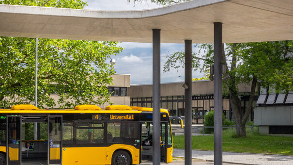 Ein gelber Linienbus mit dem Ziel "Universität" steht vor dem Sammelgebäude der Universität Regensburg