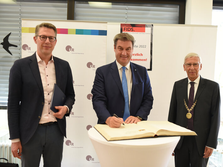 Drei Männer stehen nebeneinander. Der Mann in der Mitte hält einen Stift in der Hand und ist dabei in ein großformatiges Buch zu schreiben. (öffnet Vergrößerung des Bildes)