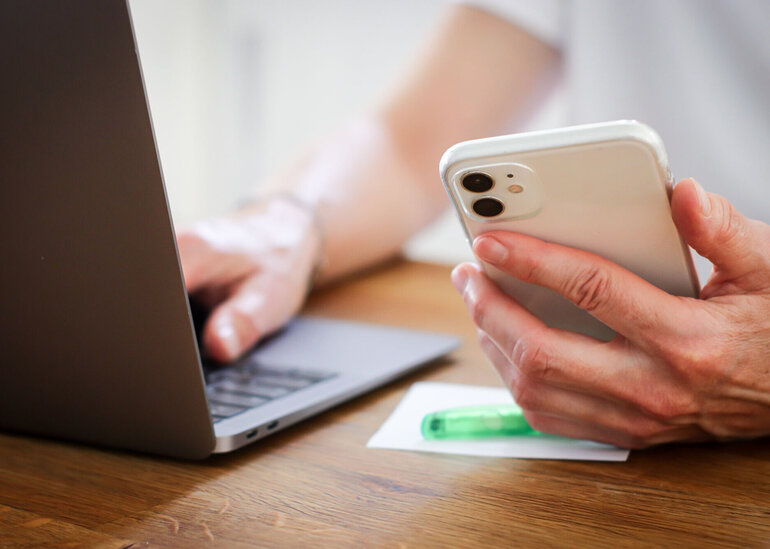 Eine Person hält ein Smartphone in der Hand und tippt etwas am Laptop.