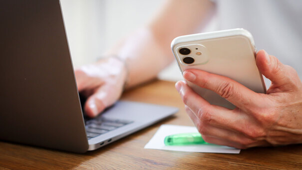 Eine Person hält ein Smartphone in der Hand und tippt etwas am Laptop.