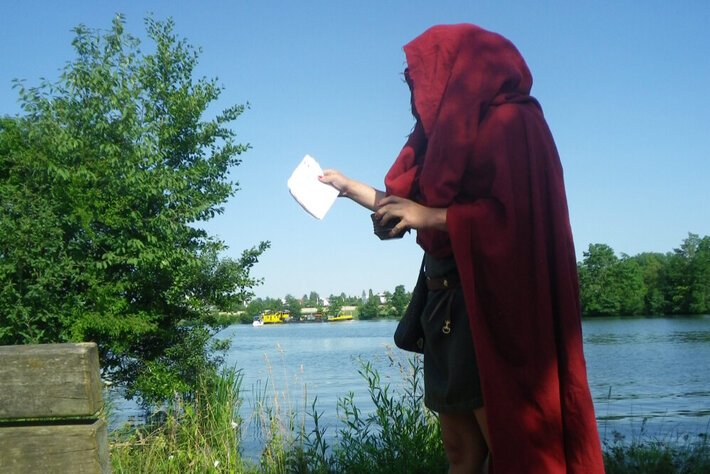 Eine Person, eingehüllt in einen roten Kapuzenumhang steht am Flussufer. In der linken Hand hält sie einen Becher, in der rechten Hand ein Blatt Papier. (öffnet Vergrößerung des Bildes)
