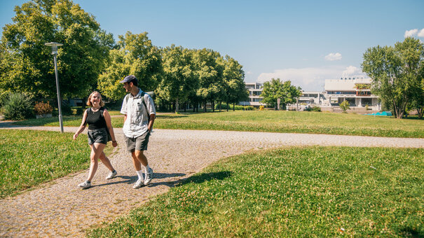 Zwei Studierende gehen im Sonnenschein, bei blauem Himmel und zwischen grünen Wiesen und Bäumen über den Campus der Universität Regensburg.