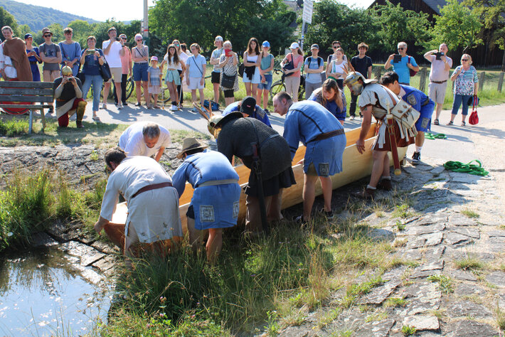 Eine Gruppe von Personen in antiken Kostümen schiebt einen Einbaum vom Ufer in einen Fluss. Im Hintergrund stehen Zuschauerinnen und Zuschauer, manche von ihnen fotografieren oder filmen. (öffnet Vergrößerung des Bildes)