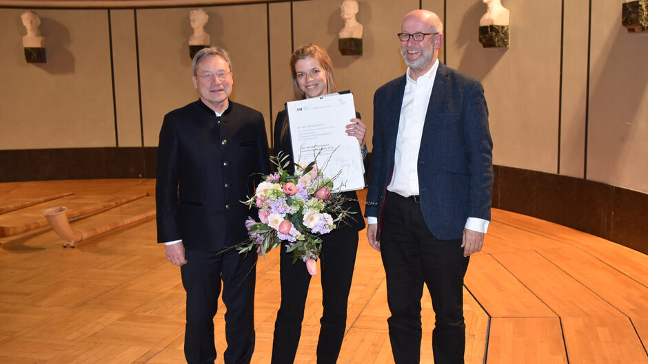 Gruppenbild mit zwei Männern, die ein lächelnde junge Frau flankieren, die einen Blumenstrauß und eine Urkunde in die Kamera hält.