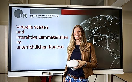 Frau Martina Rasp mit einer virtuellen Brille in der Hand vor einem Screen mit dem Titel "Virtuelle Welten und interaktive Lernmaterialien im unterrichtlichen Kontext".