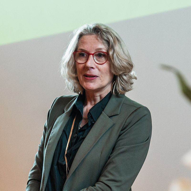 Eine Frau mit grauen Haaren und Brille mit rotem Brillengestell spricht.