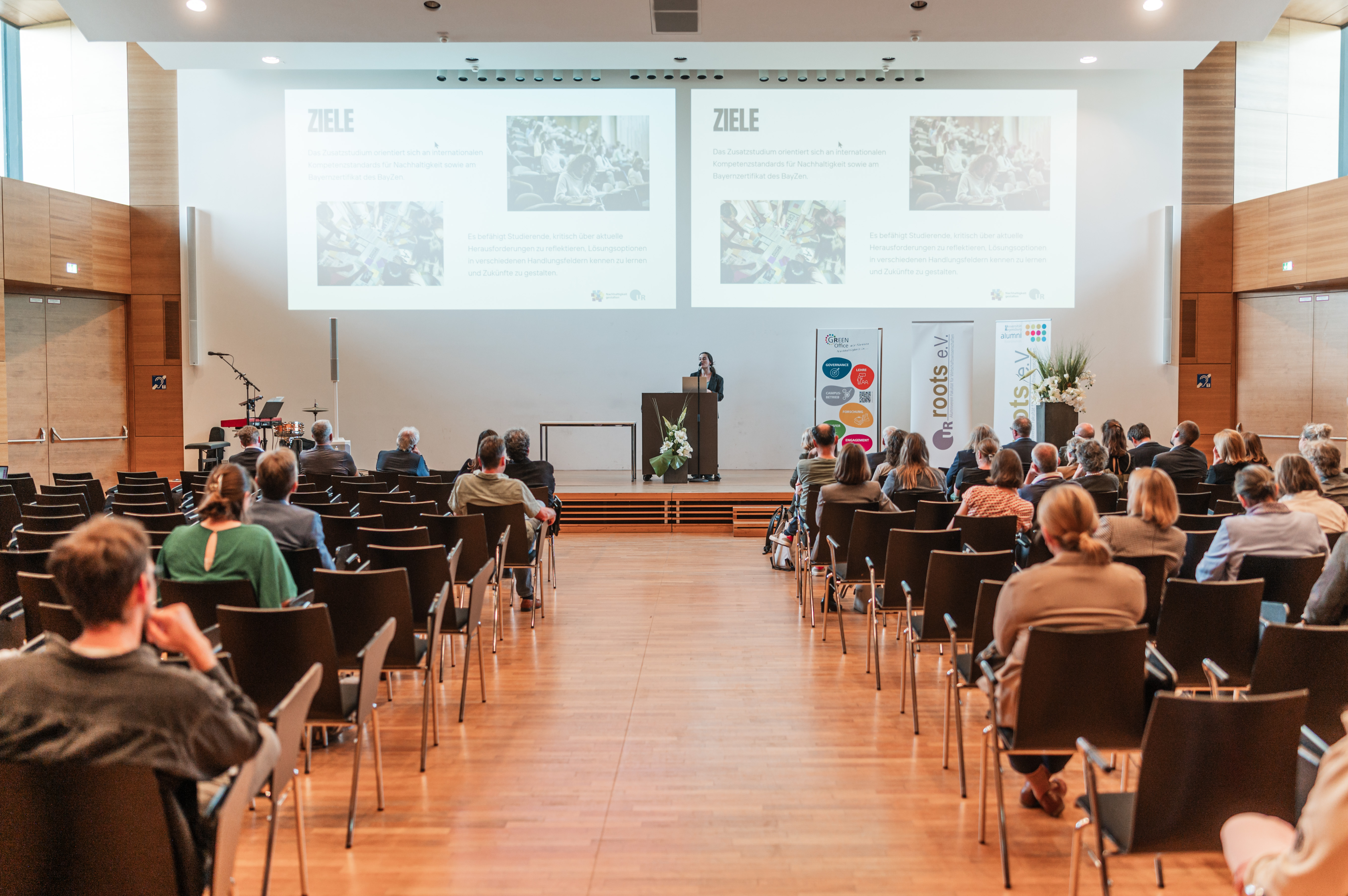 Eine Frau steht vor Publikum an einem Rednerpult. Im Hintergrund sind Folien an die Wand projiziert, auf denen "Ziele" steht. (öffnet Vergrößerung des Bildes)