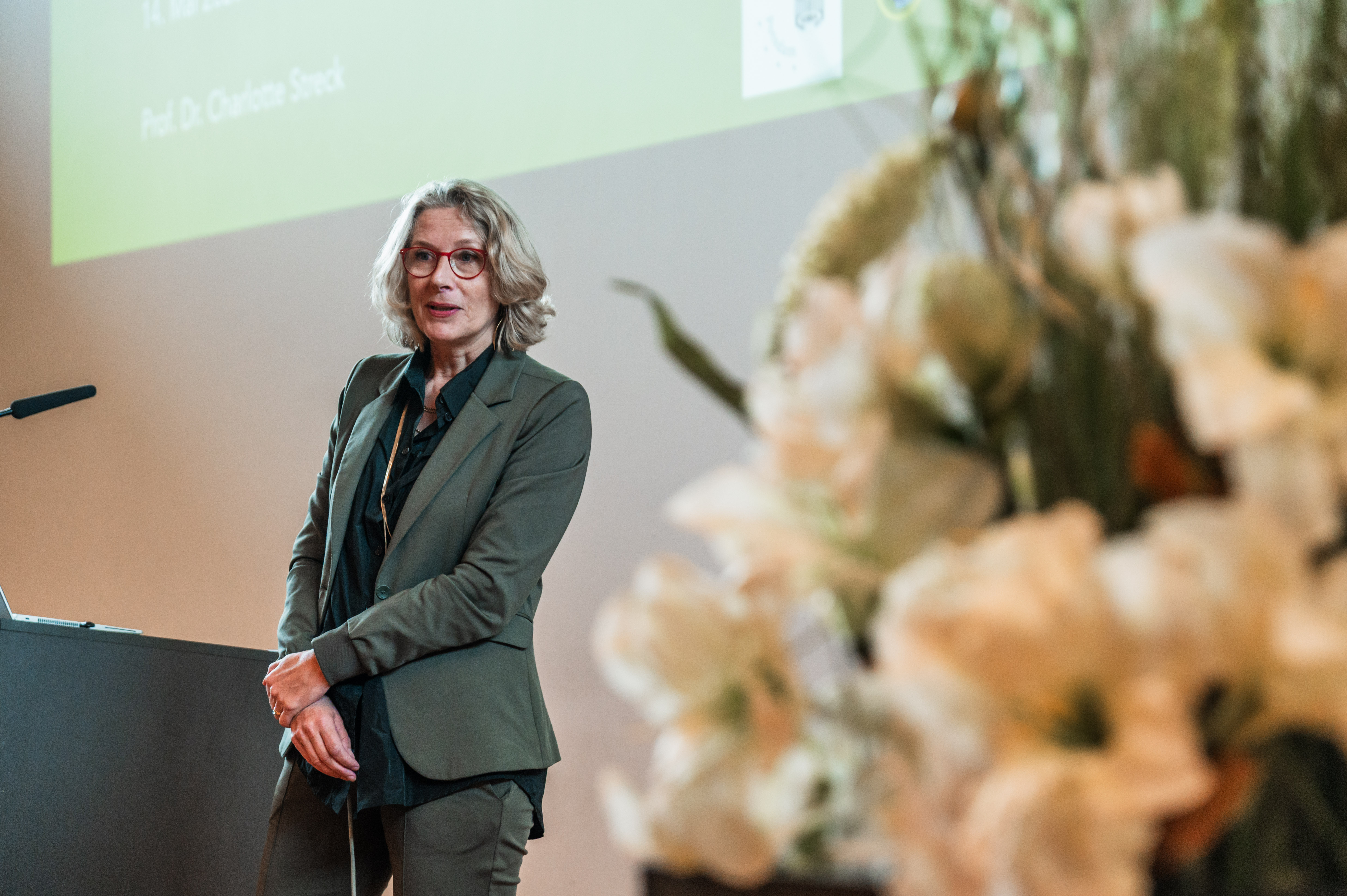 Eine Frau mit halblangen Haaren, einer Brille und einem Anzug steht auf einer Bühne und spricht. Im Vordergrund ist weißer Blumenschmuck zu sehen. (öffnet Vergrößerung des Bildes)